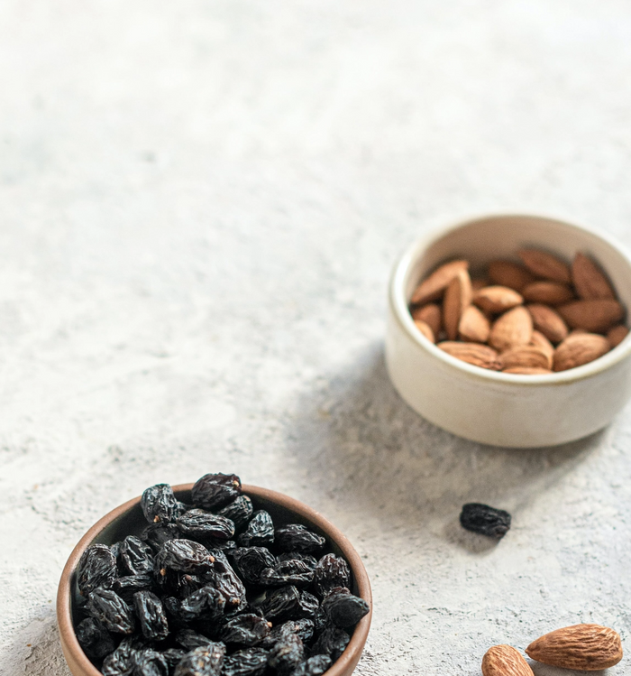Bowl of dried black raisins and a bowl of almonds on a light textured surface.